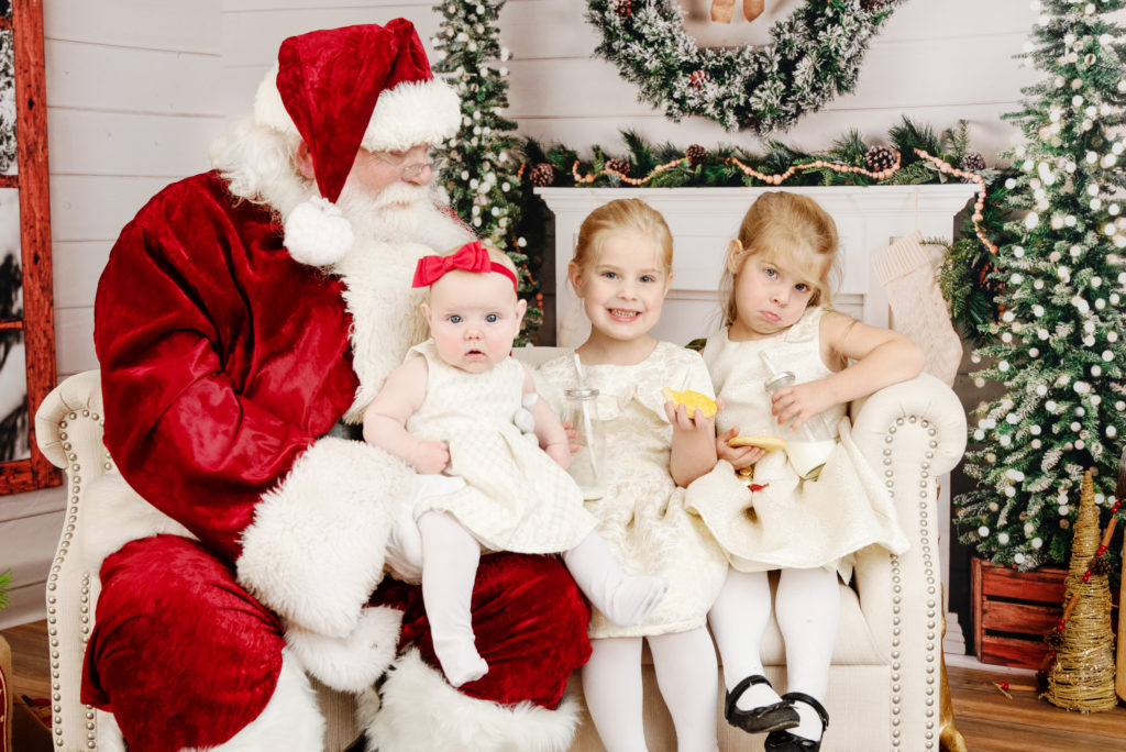 st-louis-santa-experience-santa-sitting-on-couch-with-three-little-girls-in-neutral-dresses-eating-cookies-and-milk